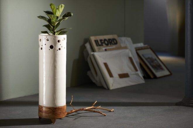 A thin, tall, off-white planter pot with a wood base and holes toward the top of the vase. A green plant sticks out from the top, and a brown root emerges from the bottom.