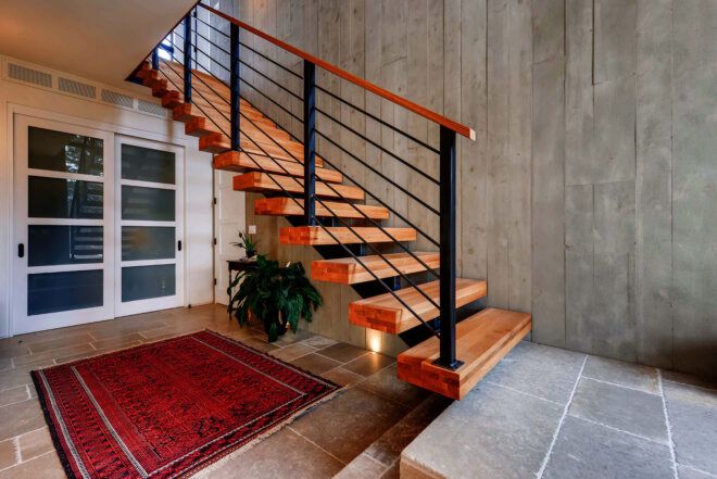 An entryway with a red rug, green potted plant and a wood staircase to the left.