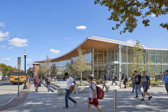 People milling around in front of a building with a wood-accented roof and windowed facade.