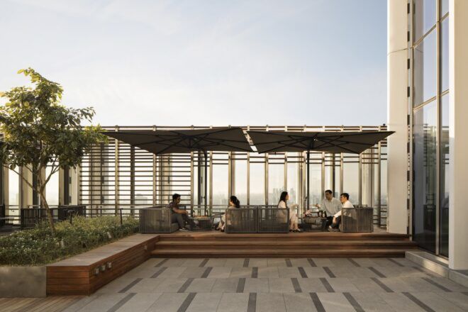 A plaza area with wood benches and a covered sitting area where people lounge. To the left there are small trees and shrubs.