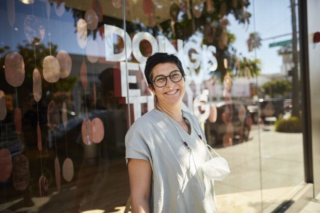 Tanya Aguiñiga standing in front of a window with her "doing is everything" artwork display.