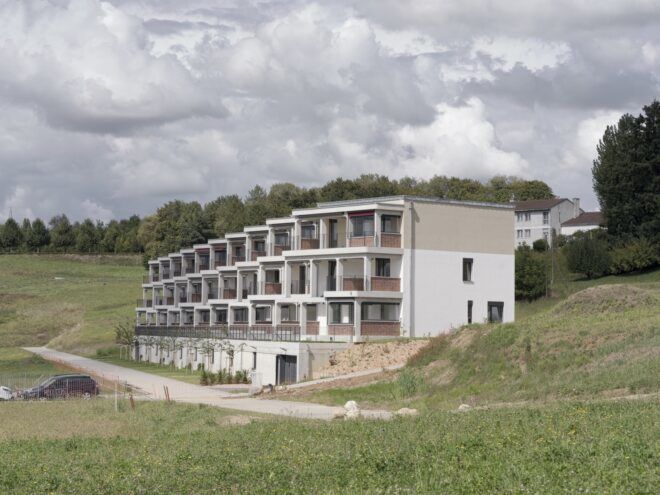 A rolling green landscape with a housing complex with a vertically tiered, off-white facade.