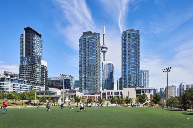 A green lawn with people walking on it. In the background, there are skyscrapers.