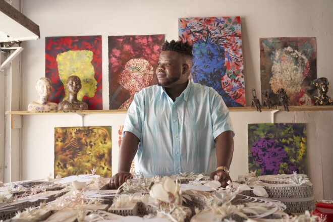 Morel Doucet standing in a studio with various artworks on the wall behind him.