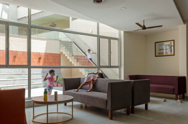 Children lounging on a couch in a room with a glass wall that looks into a building with a staircase.