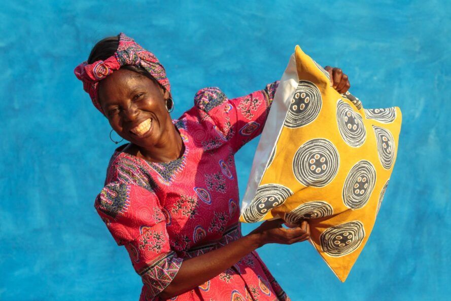 A person in a pink dress and headwrap holding a yellow patterned pillow in front of a blue wall.