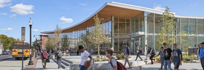 People milling around in front of a building with a wood-accented roof and windowed facade.