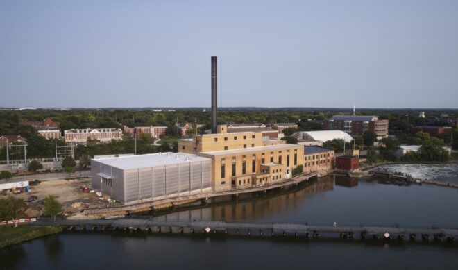 A multi-building front with facades of yellow paint and polycarbonate. A tall smokestack chimney sticks out from the roof. In front of the building is a river.