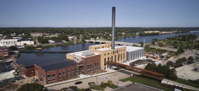 A multi-building front with facades of brick, yellow paint and polycarbonate. A tall smokestack chimney sticks out from the roof. Behind the building is a river.