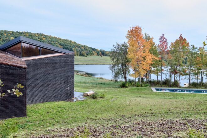 To the left, a corner of the home. To the right, trees and an expansive green lawn by a lake.