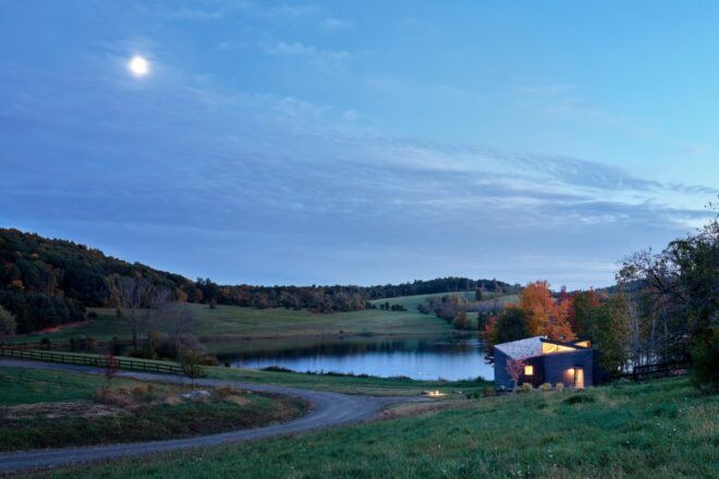A landscape at dusk, showing a curving lane by a small lakeside home lit up from within.