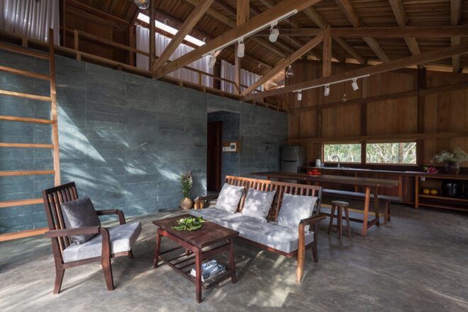 A living room with blue tile walls and a wood sofa and chair with light-colored cushions. A wood coffee table sits between the chair and sofa. In the background is a kitchen.