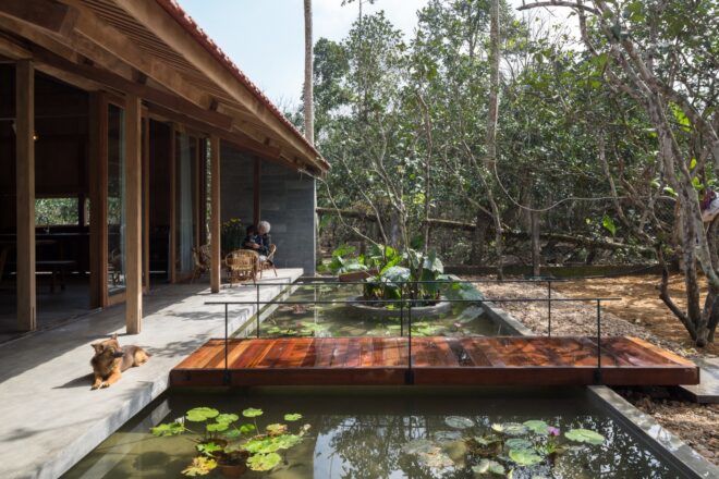 A small wood bridge crossing over the pond in front of the home. On the left, a dog lounges on the patio.