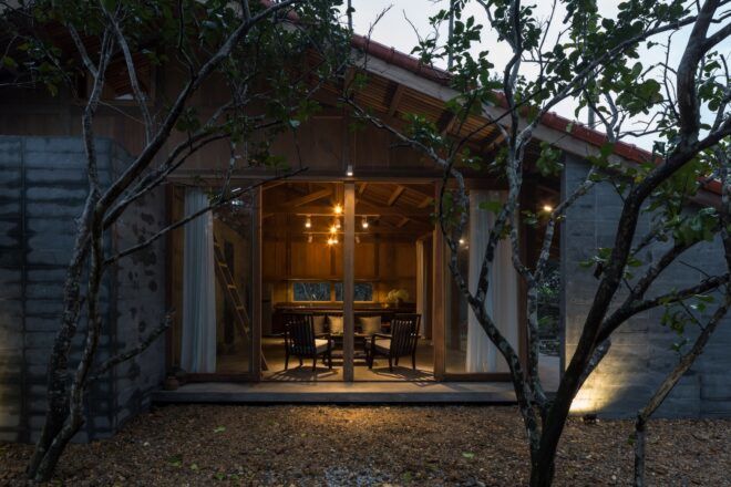 Trees outdoors partially obscuring a sliding glass door that looks into a sitting area with warm lighting.