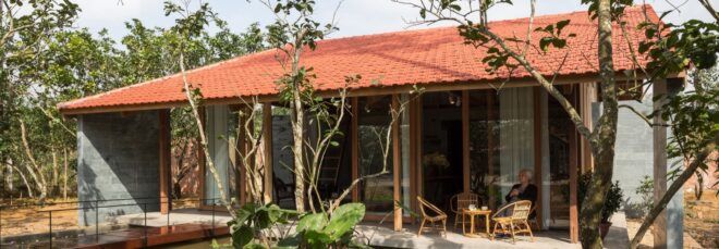 A lily pond in front of a simple home with a red gabled roof. A person sits on a chair on the patio in front of the home's sliding glass doors.