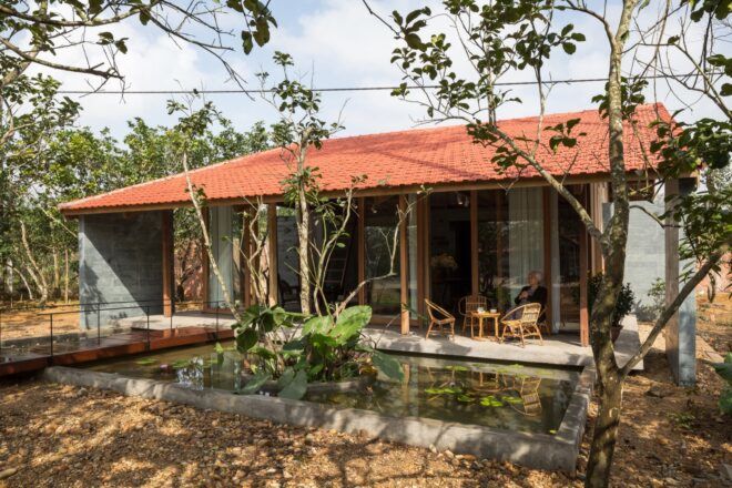 A lily pond in front of a simple home with a red gabled roof. A person sits on a chair on the patio in front of the home's sliding glass doors.