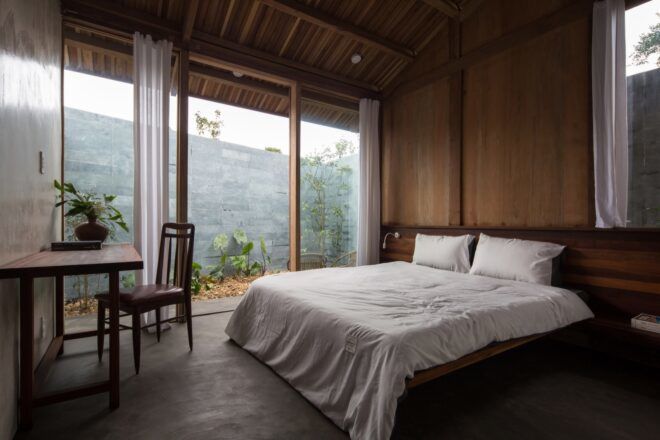 A bedroom with a simple white bed, wood walls and a desk and chair. Sliding glass doors connect to a patio area.