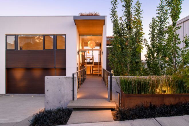 A rectangular home with a garage to the left and a walkway toward the front door at the center of the image. To the right are green plants.