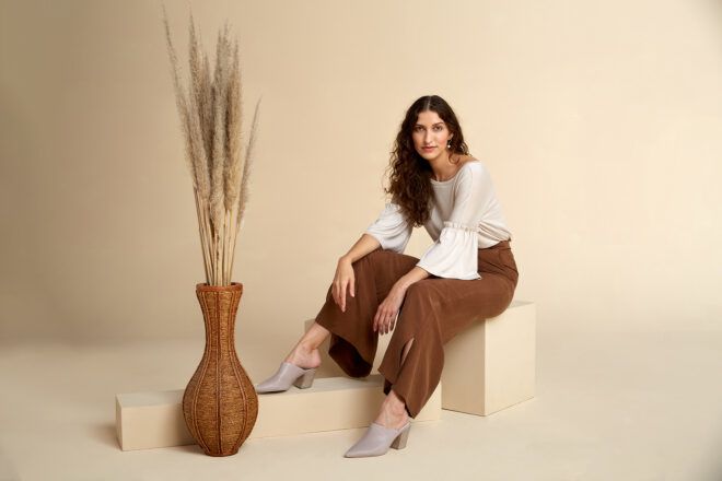 A person sitting on a beige block next to a brown pot of beige flowers. The person is wearing a white shirt and brown pants.