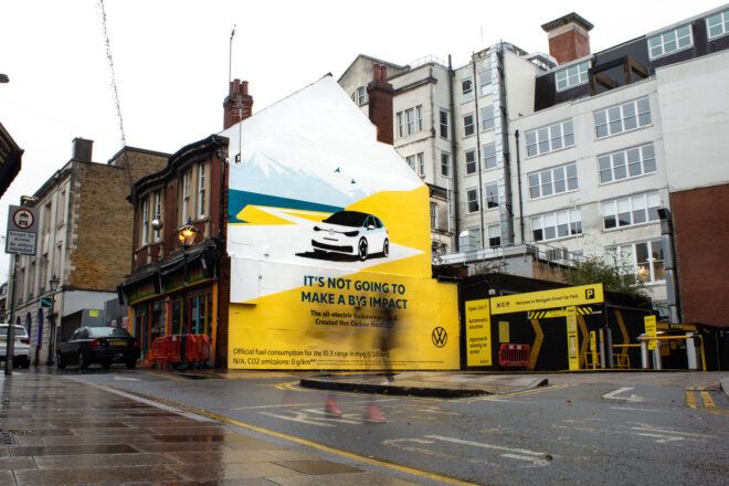 A large painted VW ad on the side of a building. A blurry figure walks past on the street in front of the building.