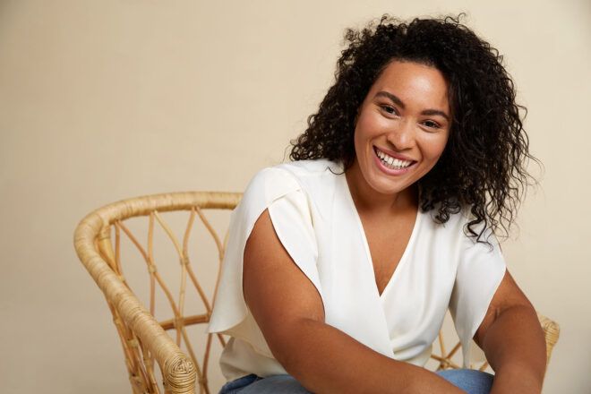 A person sitting in a wicker chair, smiling at the camera and wearing a white, short-sleeved shirt.