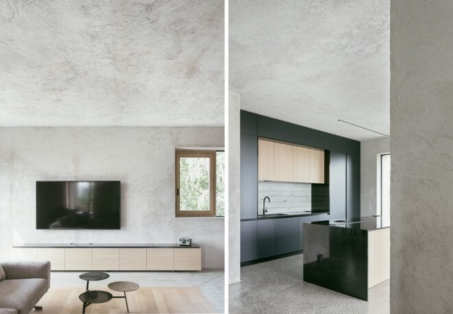 On the left, wall-mounted TV in room with raw concrete walls, floors and ceilings. On the right, kitchen with black cabinets.
