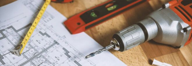 A drill and various other tools laid out on a table next to blueprints.