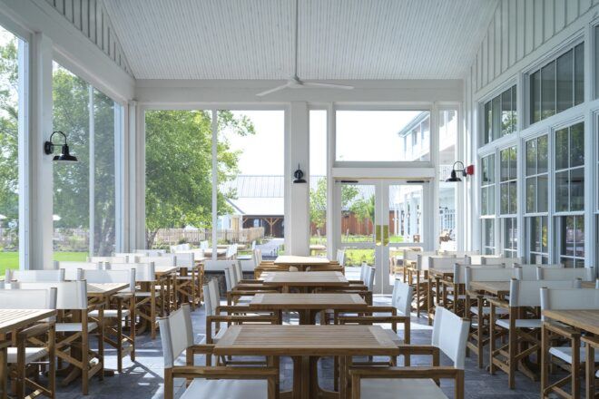 A dining room with many wood tables and chairs, and white walls with large windows.