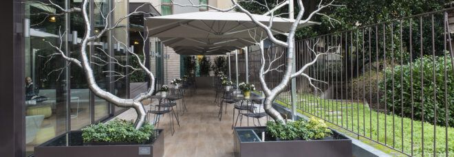 A walkway to a hotel building with white stone path, potted green bushes and two metallic tree sculptures.