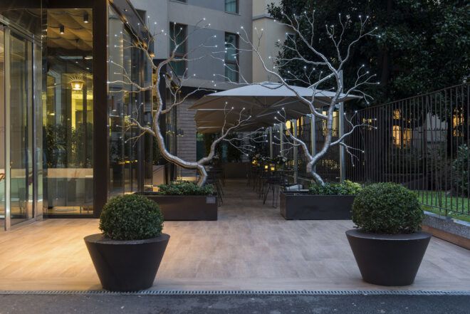 A walkway to a hotel building with white stone path, potted green bushes and two metallic tree sculptures.