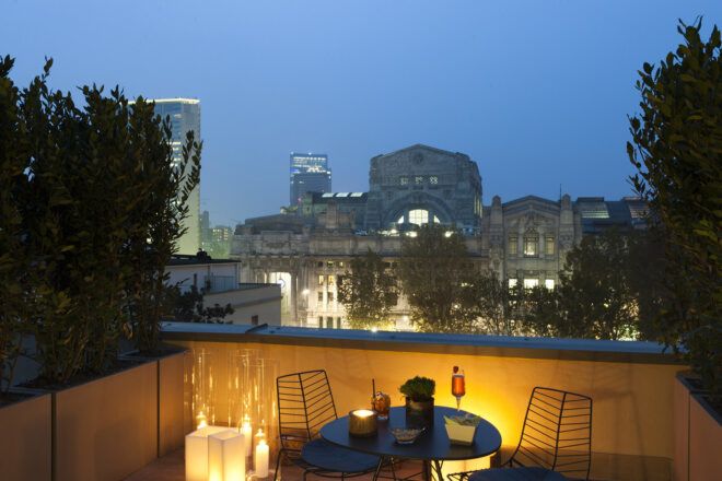 A green rooftop with a round table and two chairs that overlook Milan at night.