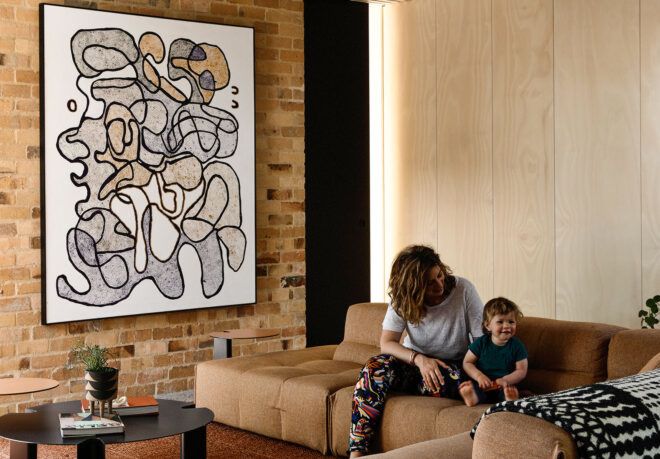 A living room with a large piece of abstract art on a beige brick wall across from a parent and child sitting on a brown sofa.