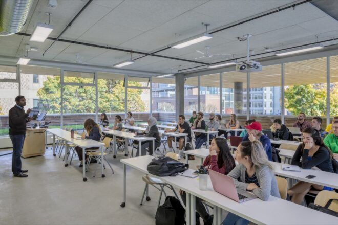 college students sitting at desks in room with large windows