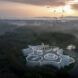 aerial view of large white building with curved walls and green roofs near a forest at sunset