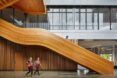 The interior of a building with a gently curving wood staircase, with two people walking in front of it.