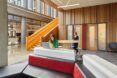 A red front desk in the lobby of a building. Two people stand behind the desk. To the left is the entrance to a wood staircase.