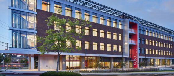 A streetview of the exterior of a multi-story building with a brick facade and a tree in front of it.