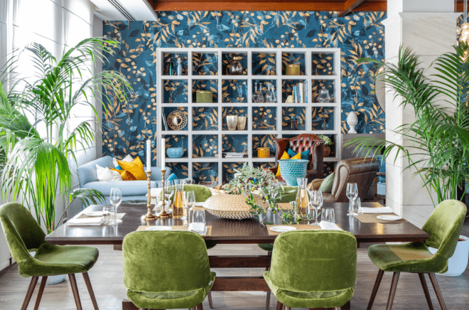 wood dining table and green velvet chairs facing blue and green floral wallpaper