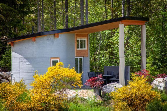 A curved concrete home with an overhang that covers a small outdoor patio with chairs and a table. Greenery surrounds the home.