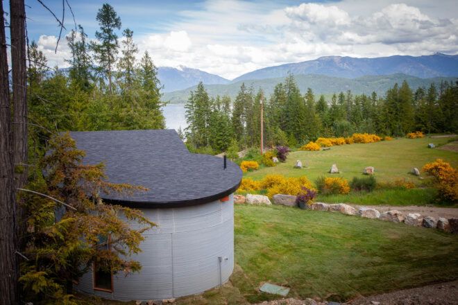 To the left, the back of a curved concrete home. To the right, a green field.