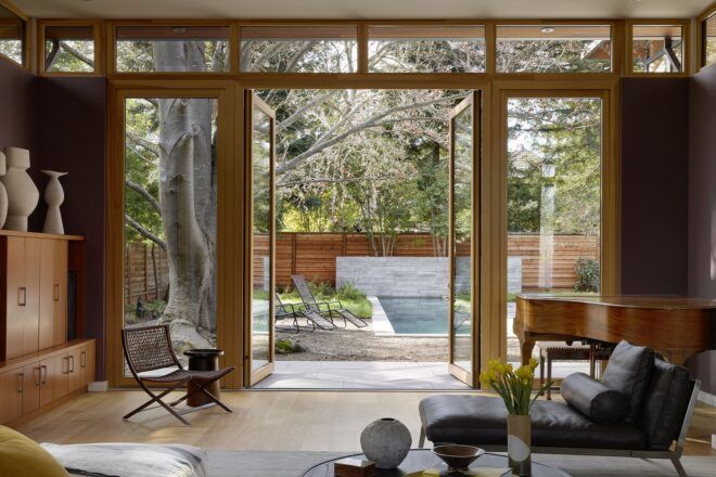 A living room with a dark gray chaise lounger that looks out onto open floor-to-ceiling glass doors that show the backyard.