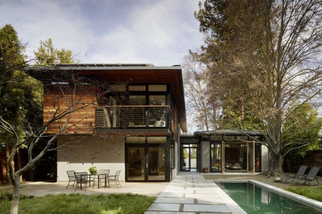 A two-story home with the top floor featuring wood siding. A third, box-like structure is attached to the lower story. A pool and patio sit in the backyard.