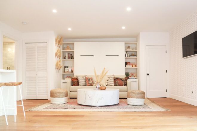 A bright living room with white walls and light-colored wood flooring. A cream colored sofa sits in front of two bookshelves.