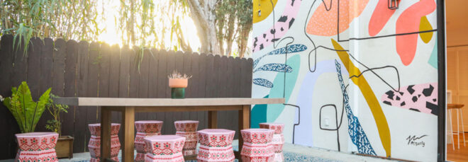 An outdoor patio space with a colorful wall painted with abstract shapes and a dining table with several pink stools around it.