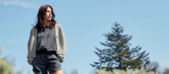 A person wearing a shirt, cardigan and shorts stands on the left, looking off into nature.