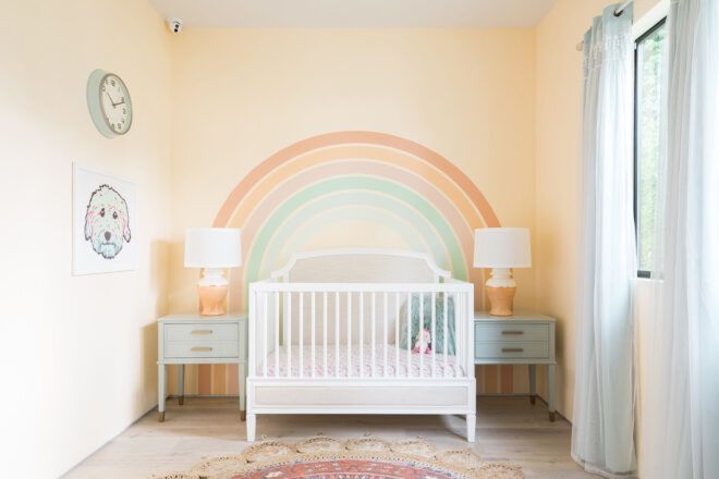 A nursery with a white crib, two cribside tables, two lamps and a yellow wall featuring a painted on rainbow.