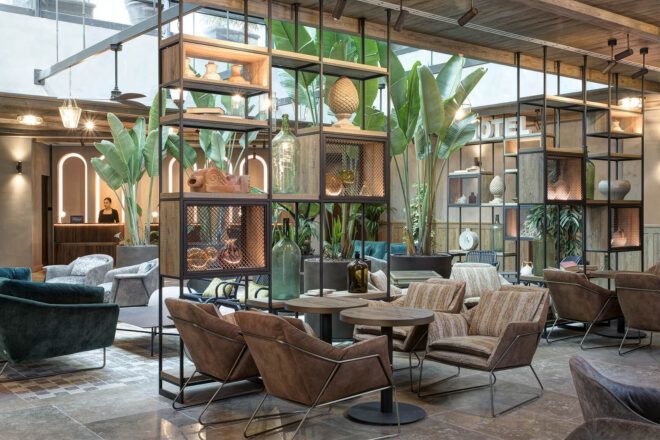 Two tables are surrounded by four cream colored chairs in a sitting/dining area with open bookshelves separating the sitting area from a courtyard with plants.