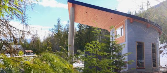 A curved concrete home with an overhang that covers a small outdoor patio with chairs and a table. Greenery surrounds the home.