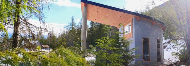 A curved concrete home with an overhang that covers a small outdoor patio with chairs and a table. Greenery surrounds the home.