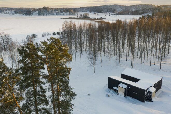 snow-covered black cabin in forest facing a lake